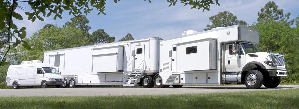 Three white mobile trailers and vehicles parked in a row on grass with trees in the background.